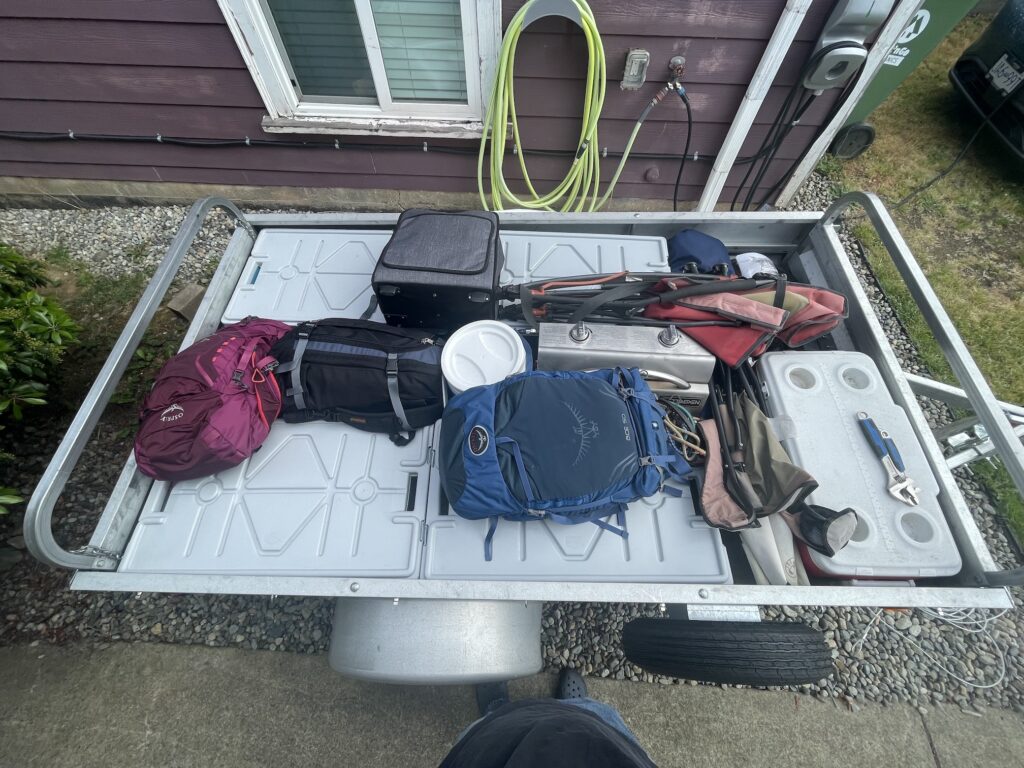 An overhead picture of the packed trailer shows for grey cover topped totes and a number of backpacks overtop of them. Also a cooler is in the right front corner and camping chairs and stove are in the spaces between totes.