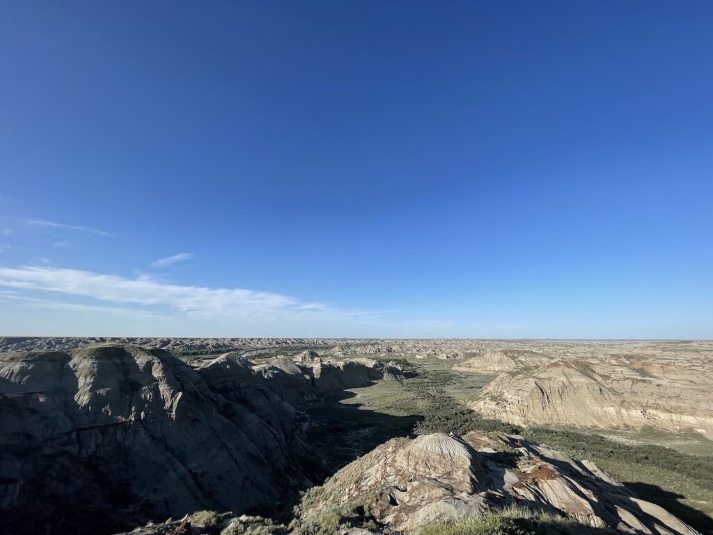 A huge blue sky with a line of white clouds in the distance on the left. The horizon is perfectly flat. Below the horizon is the badlands, brown irregularly shaped hills and canyons stretch for as far as the eye can see.