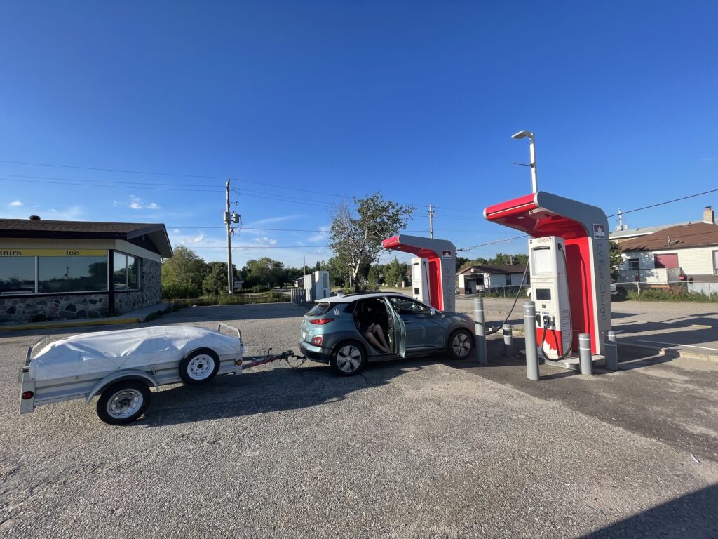 The car and trailer are seen parked straight into two red and white Petro Canada charging stations. The sky is a clear blue above. There is a small building behind to the left of the gas station. To the right behind the chargers is a small road and a home.