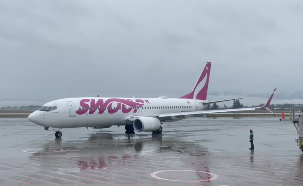 A Swoop Airlines 737 is pictured on a wet tarmac in Abbotsford. There is a person standing under one wing. The plane is sideways to the camera, nose to the left.