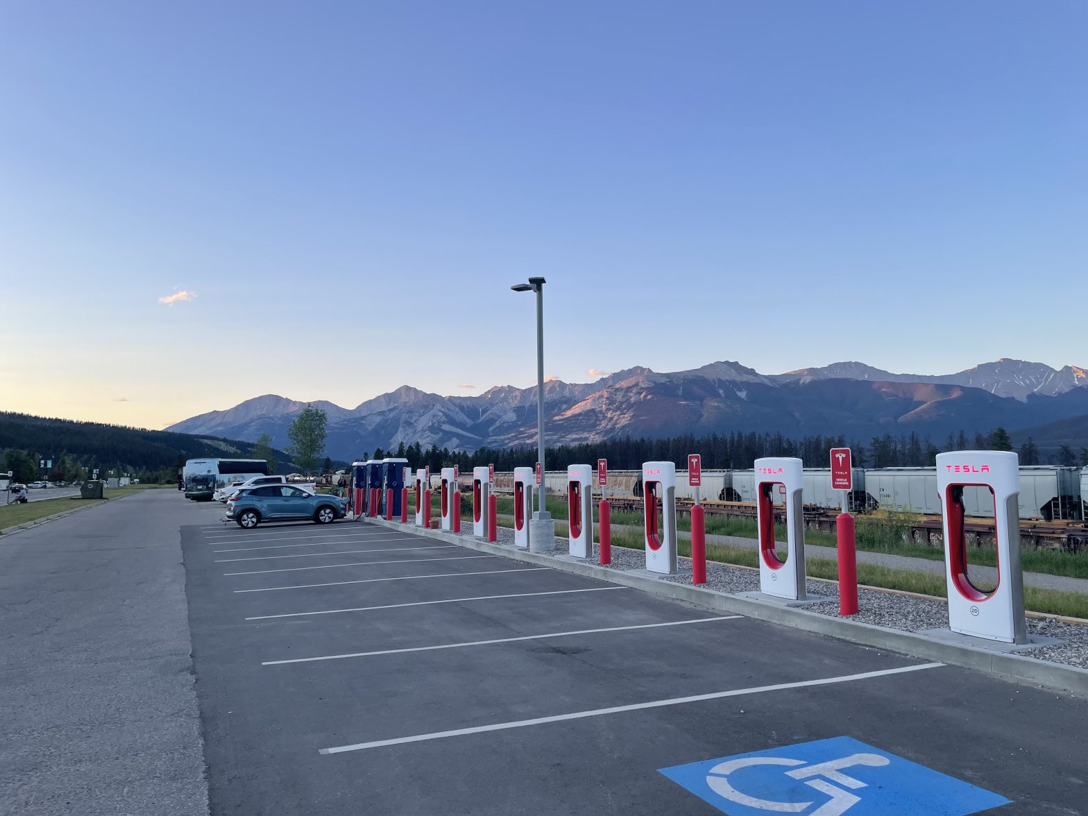 A sunset shot shows a long parking lot with a long line of Tesla superchargers. In the distance, perhaps 5 spots down, is a blue Hyundai Kona. This is Jasper, charging beside the railway.